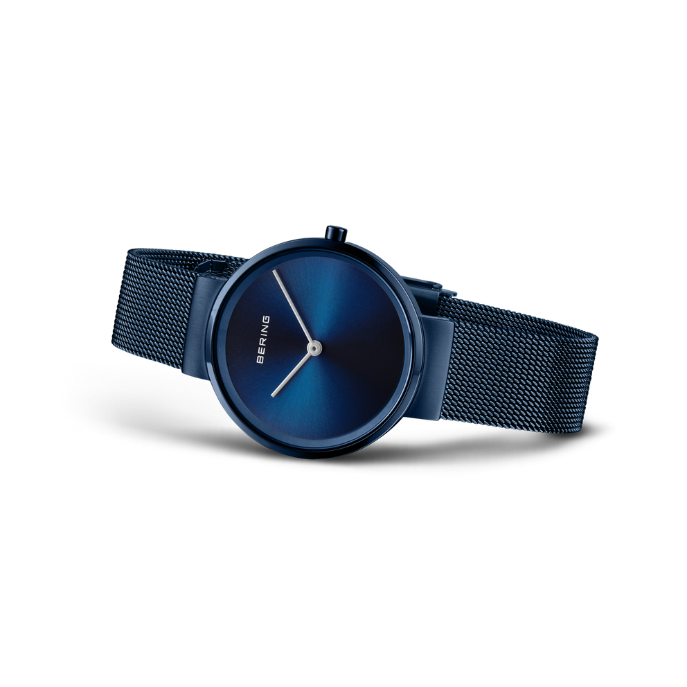 Reloj de pulsera con esfera azul redonda, manecillas delgadas, pulsera de malla oscura, vista en ángulo.