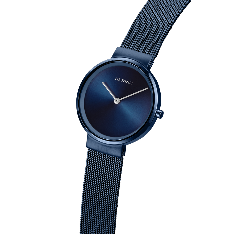 Reloj minimalista azul oscuro con correa de malla, vista lateral mostrando esfera y detalle de correa texturizada.