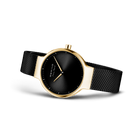 Wristwatch with black dial, white hands, gold case, and black mesh band, shown at angle.