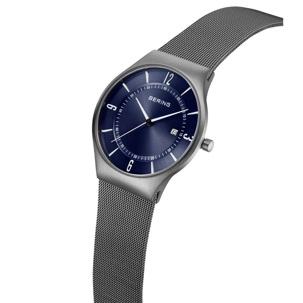 Reloj de pulsera con esfera azul oscuro, agujas e índices plateados, y pulsera de malla gris de acero, en vista de ángulo.