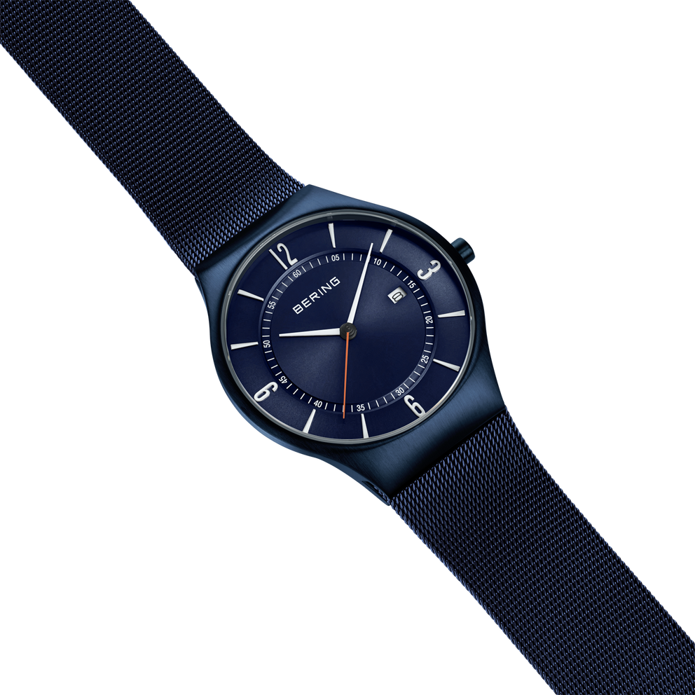 BERING wristwatch with dark blue dial and black case, displayed at angle with dark blue mesh strap against light blue background.