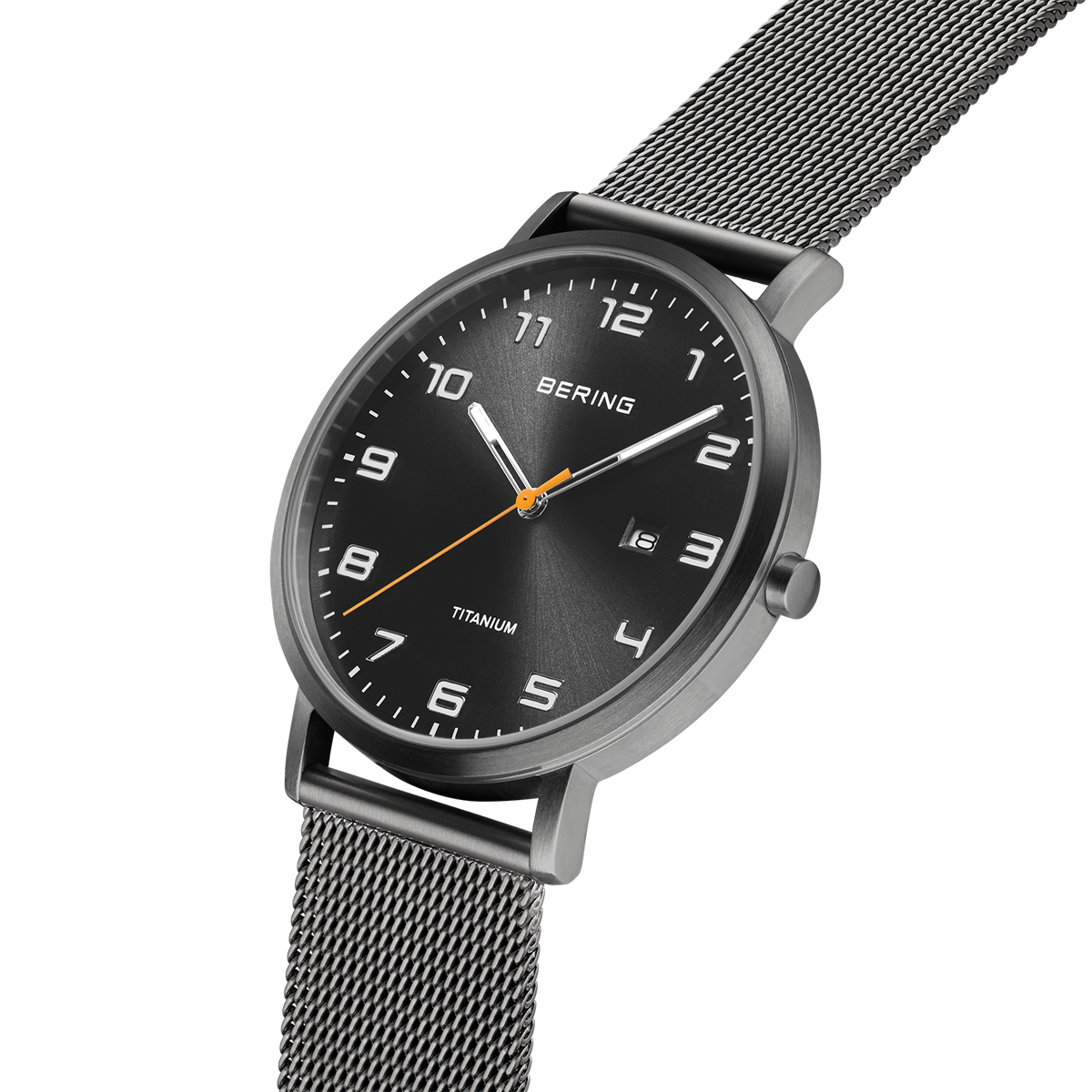 Black-dial BERING titanium watch with white hour markers, white and orange hands, shown at angle with mesh metal bracelet.